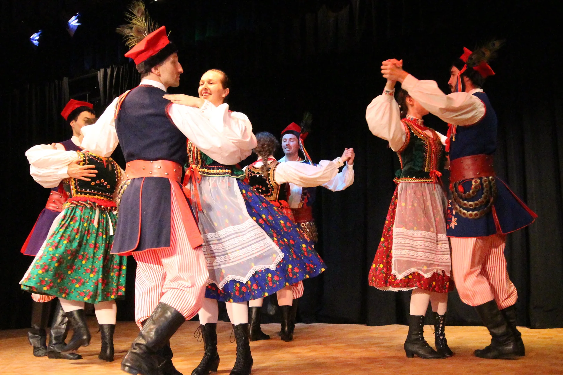 Dancers in traditional Kraków folk costume — peacock-feather caps, red aprons and black boots — mid-dance on a lit stage